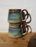 Two stacked ceramic mugs with a blue, green, and brown speckled glaze, resting on a wooden surface against a white wall.