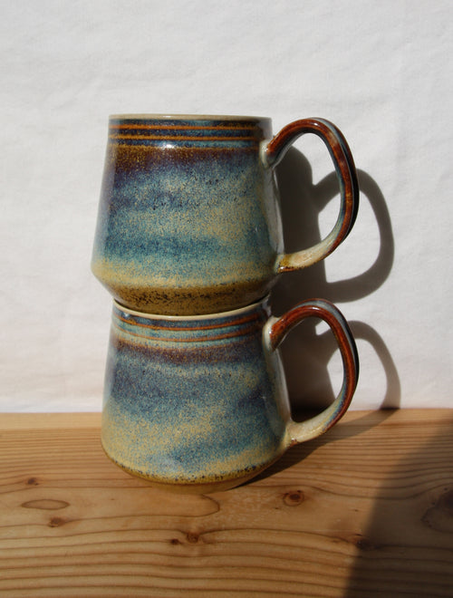 Two stacked ceramic mugs with a blue, green, and brown speckled glaze, resting on a wooden surface against a white wall.